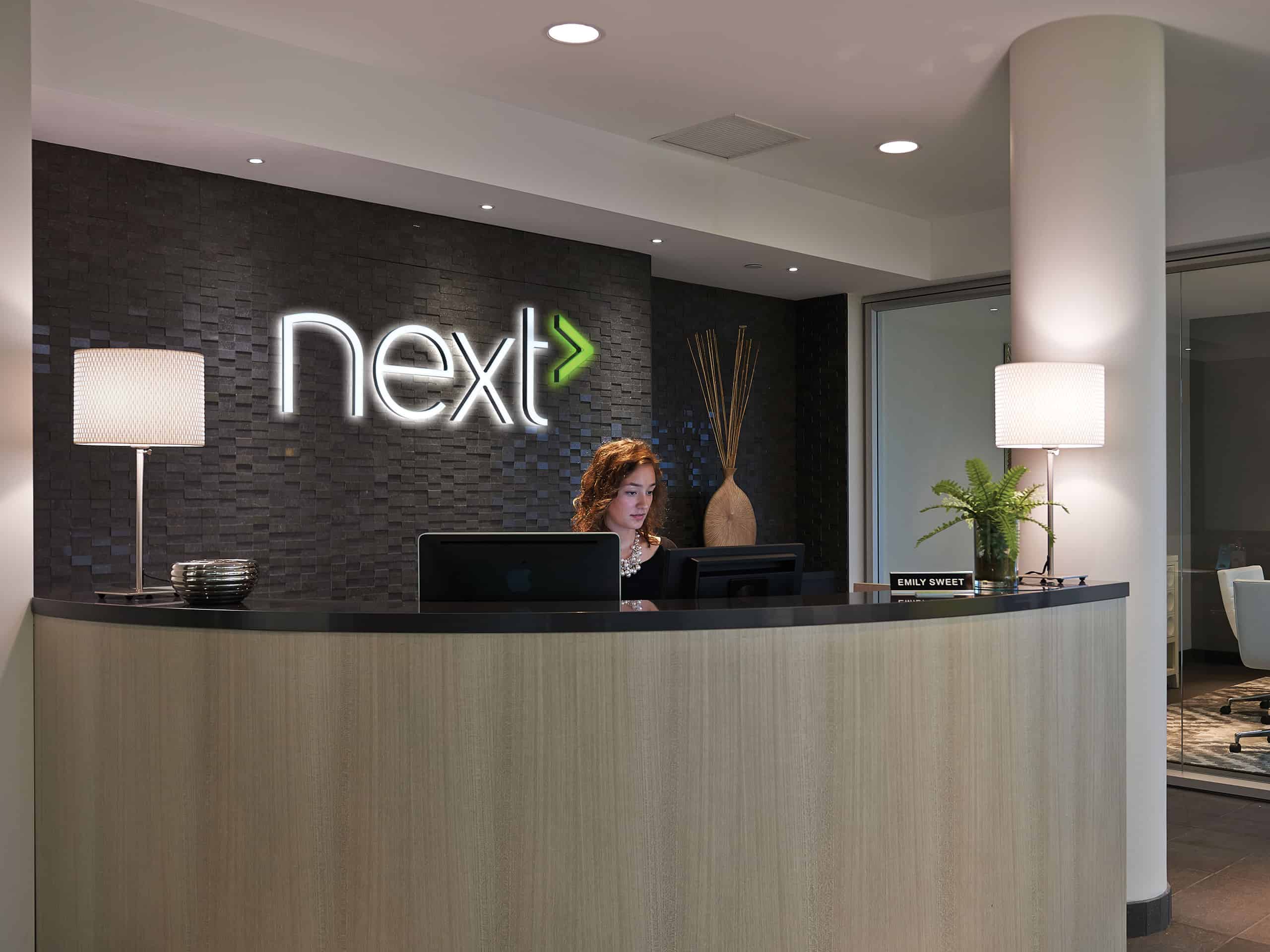 A woman is standing at the front desk of a next hotel featuring custom signage behind her desk.